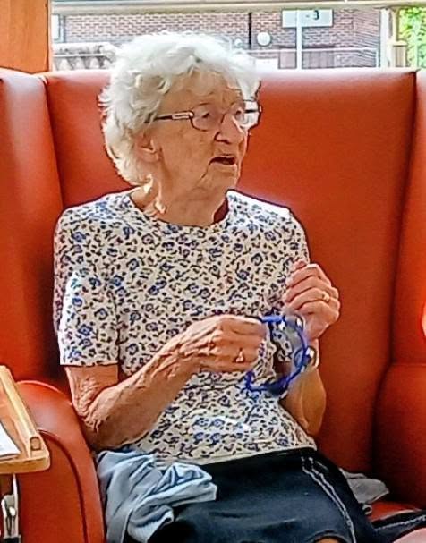 Elderly woman with grey hair and spectacles, wearing a patterned top and seated in an orange chair, holds a pair of blue-framed spectacles, ready to enjoy a Two Can Uke session.