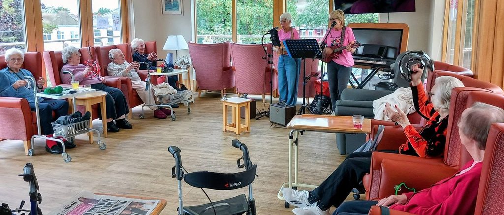 The duo Two Can Uke perform with microphones and guitars for a group of elderly people seated in a brightly lit common room with large windows, armchairs, and mobility aids.