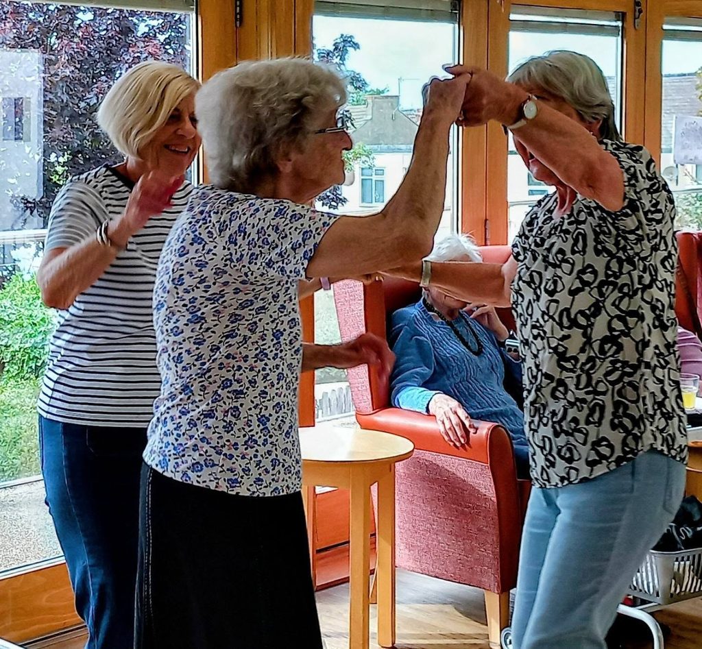 Two elderly women dance together whilst another woman claps and smiles nearby in a bright room with large windows, as Two Can Uke music plays. People are seated in the background.