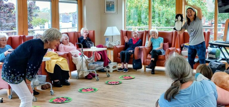 A group of elderly women sit in armchairs, watching a woman standing and holding up a puppet. Another woman leans towards them, while children sit on the floor facing the group in a bright room with large windows.