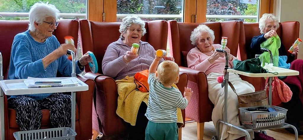 During Music Time, a young child stands before four elderly women holding colourful shakers in a bright community room, creating a joyful intergenerational session.