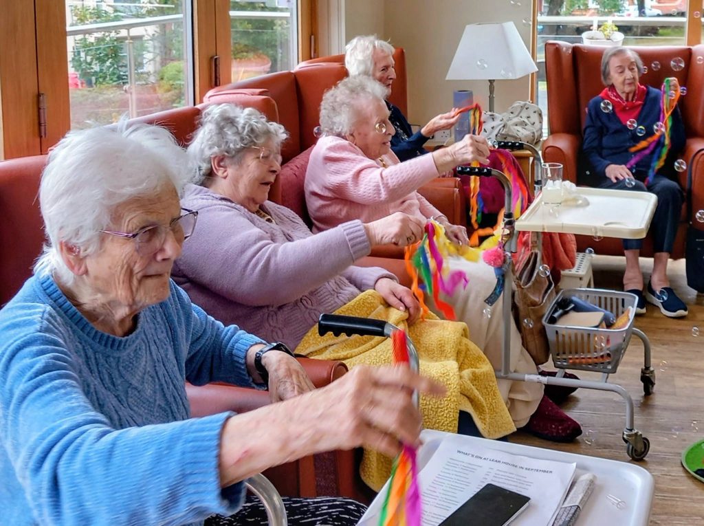 A group of elderly women sit in armchairs in a brightly lit room, smiling and waving colourful ribbons during Music Time, with mobility aids and small tables nearby.