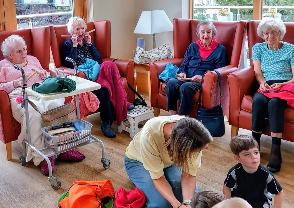 Five elderly women sit in armchairs in a bright room during an intergenerational session, whilst a woman kneels to assist a young boy. Other children and bags are scattered across the floor around them.