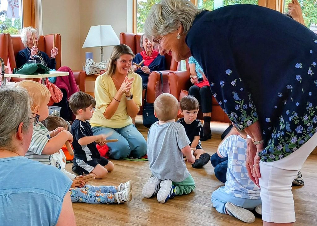 A group of children sit in a circle, interacting with an older woman during an intergenerational session. Adults observe and clap in a bright room with large windows—it’s Popcat’s Music Time!.