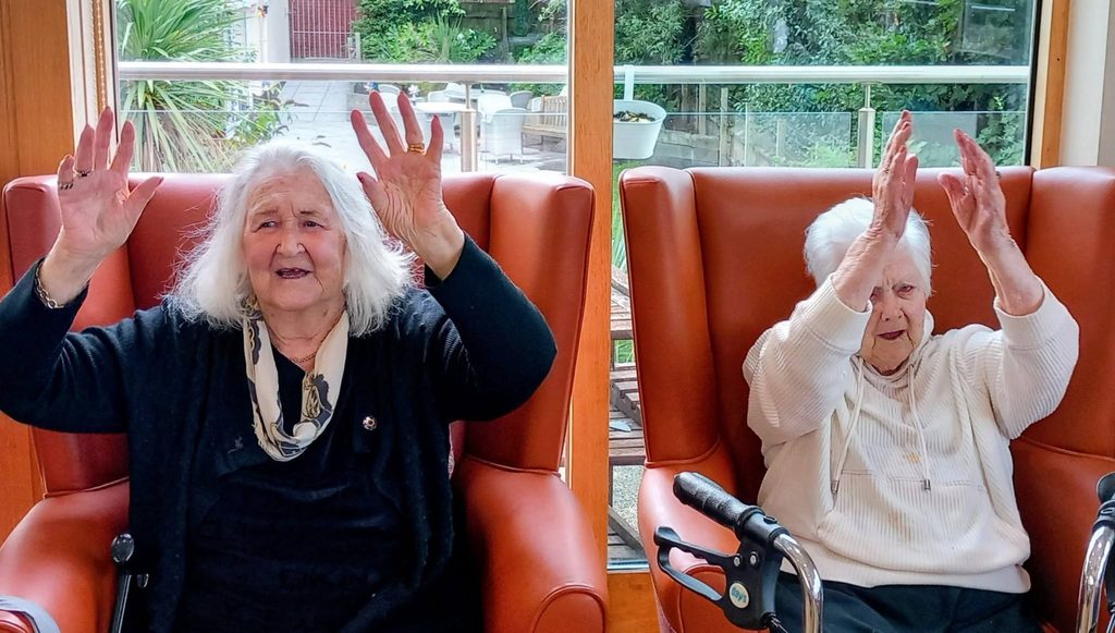 Two elderly women sit indoors on orange chairs, smiling and raising their hands during a Music Time session. A walking frame is in front of one, and large windows behind reveal verdant greenery outside.