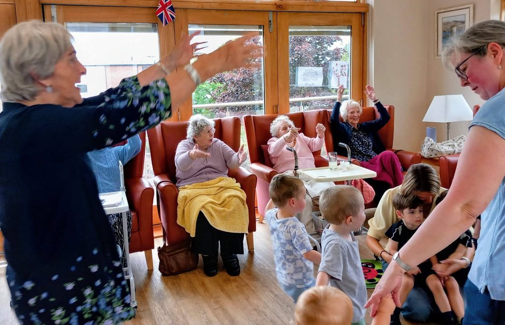 Elderly women and young children with carers enjoy an intergenerational session in a bright, communal room—sharing laughter and stories beneath a small Union Flag.