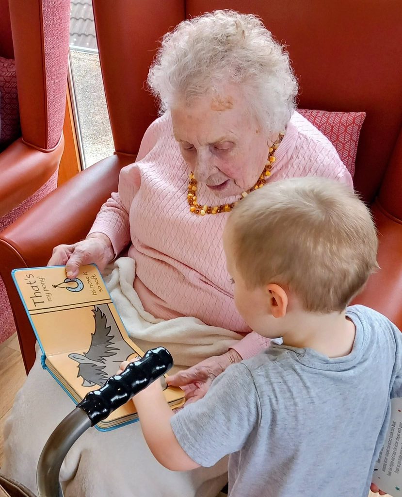 An elderly woman sits in a red chair reading a children’s book with a young boy, who is pointing at an illustration of a bird during their intergenerational session.