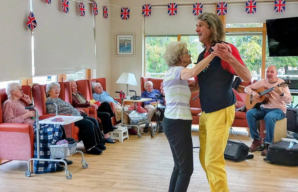 An older woman and a man with long hair dancing in a room decorated with British bunting for a Fabulous Afternoon, whilst other people sit in armchairs and one man plays an acoustic guitar.