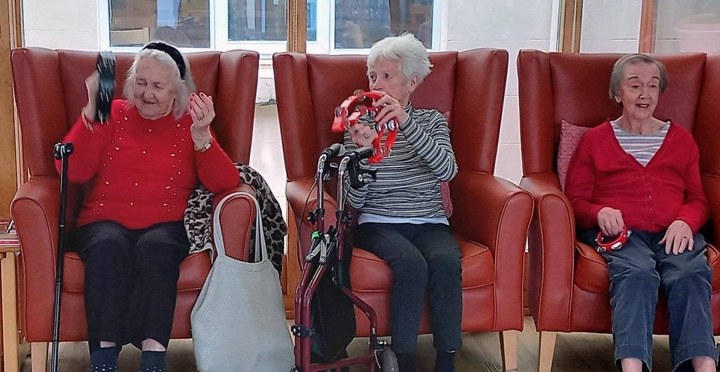 Three elderly women sitting in leather wingback chairs playing hand percussion instruments during a Fabulous Afternoon show.