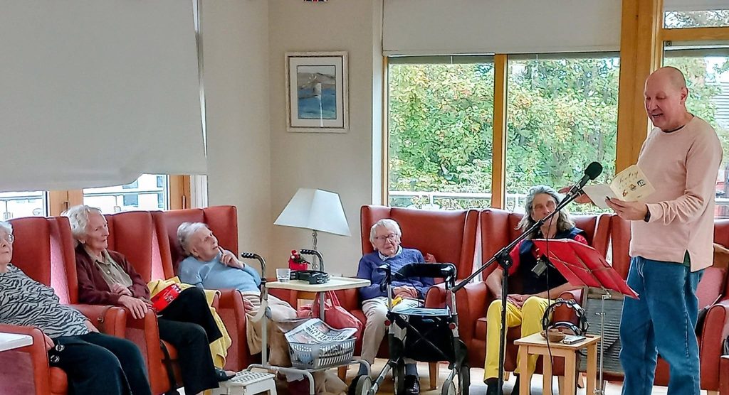 Marko reads from a pamphlet into a microphone whilst a group of elderly people sit in armchairs listening during a Fabulous Afternoon in a sunlit room.