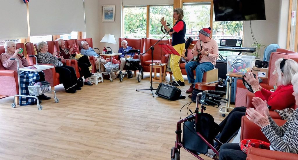 Andrew and Marko perform for a group of pensioners sitting in armchairs in a communal lounge, some of whom are holding percussion instruments.