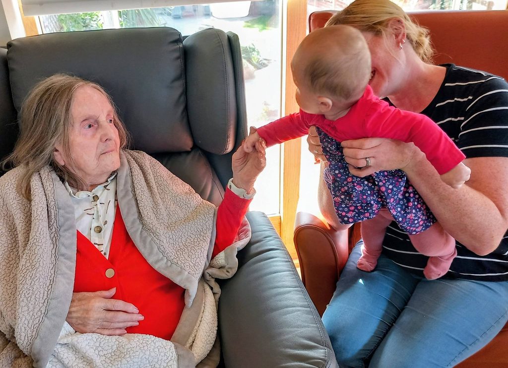An elderly woman in an armchair holds hands with a Boogie Baby, who is being cradled by a woman sitting in a chair beside her.