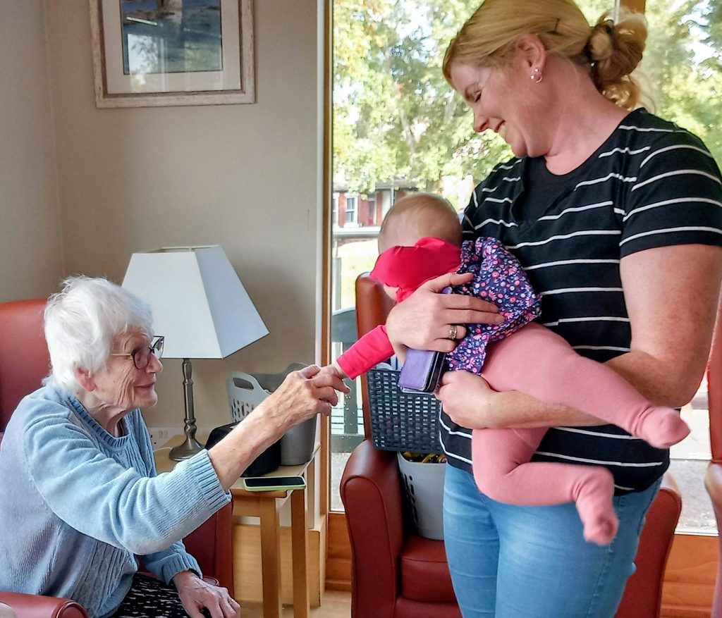 An elderly woman sits in a chair and reaches out to hold the hand of a Boogie Baby being held by a woman standing beside her.