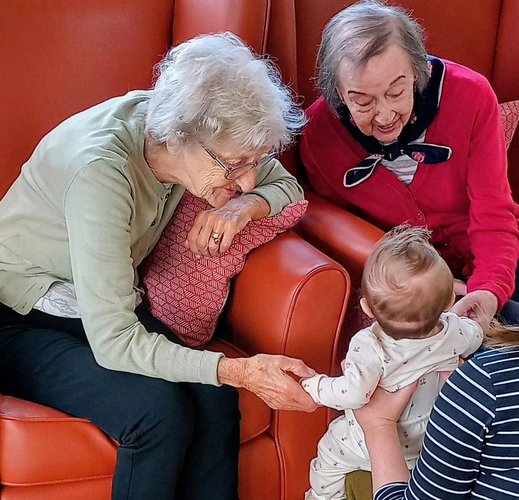 Two elderly women sitting in armchairs hold hands with a dancing baby.