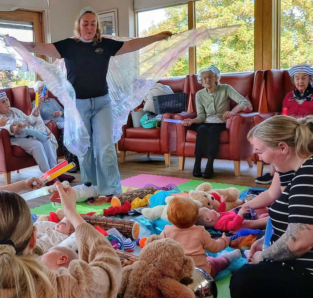A woman holds out iridescent fabric wings during a Boogie Baby session, as babies lie on floor mats surrounded by toys while older adults watch from nearby armchairs.
