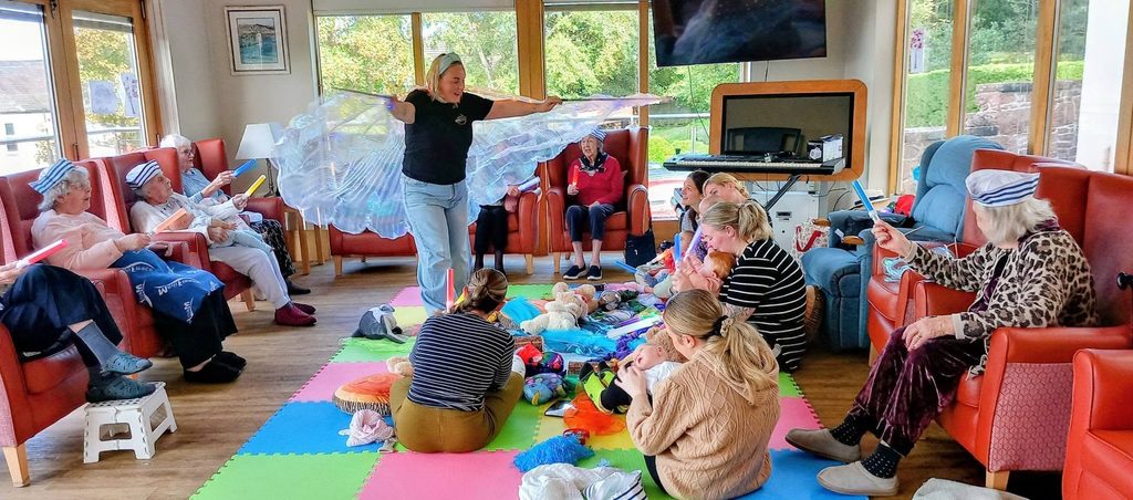 An adult leads a Boogie Baby nursery-style activity with a group of elderly residents and young children sitting on colourful floor mats.