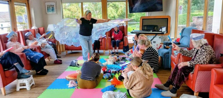 A woman holding translucent fabric wings leads a Boogie Baby activity for a group of elderly residents and young children sitting together in a care home lounge during Sailor Week.
