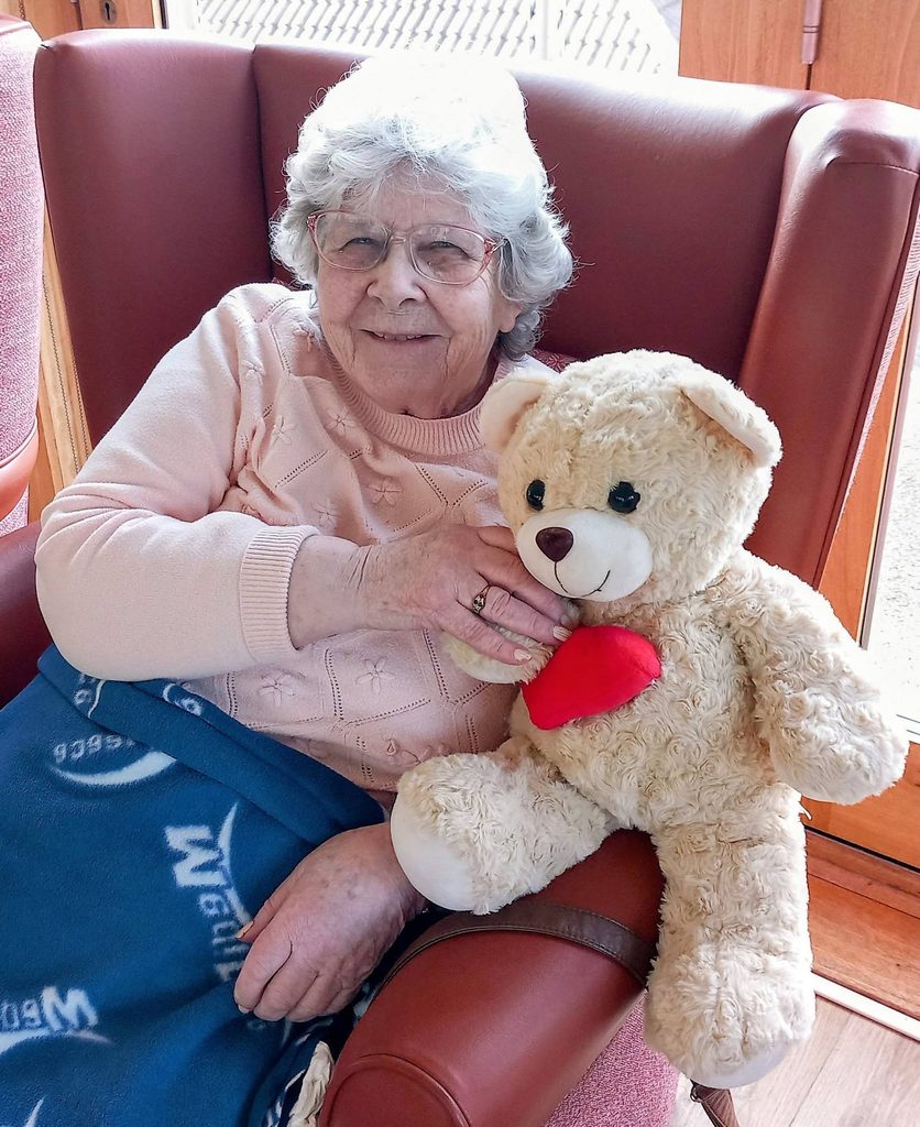 An elderly woman with grey hair and spectacles sits in a red armchair, holding a teddy bear with a red heart on its chest during a Sailor Week children’s event.