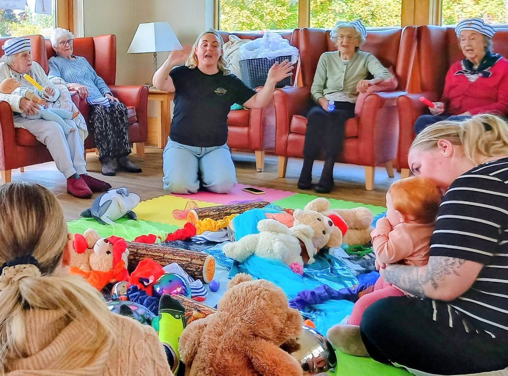 A woman leads a Boogie Baby playgroup session with elderly residents and a baby seated on a play mat, surrounded by soft toys.