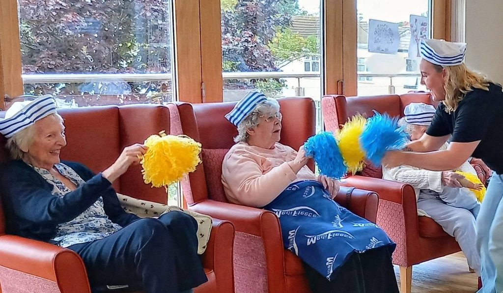 A woman in a sailor cap leads three seated older adults in a Sailor Week pom-pom activity.