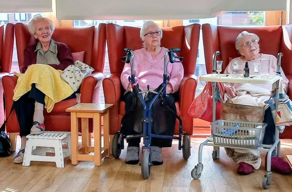 Three elderly women sit in red high-backed armchairs at Lear House; the woman in the centre uses a walking frame, and the woman on the right has a small table with spectacles attached to her walking frame.