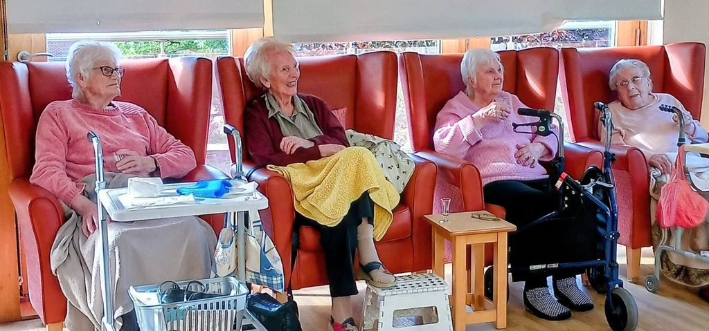 Four older women, including Jess, sit in red high-backed chairs in a Lear House communal room, with mobility aids including a walking frame and a side table present.
