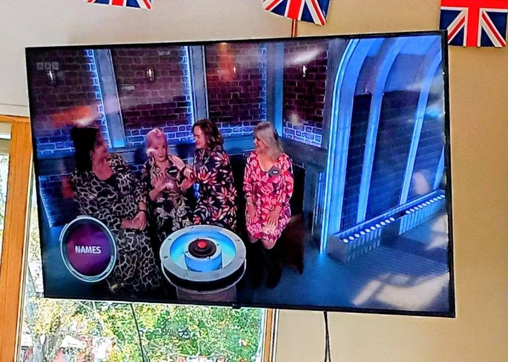Four women competing on Bridge of Lies, shown on a wall-mounted screen beneath Union Flag bunting.