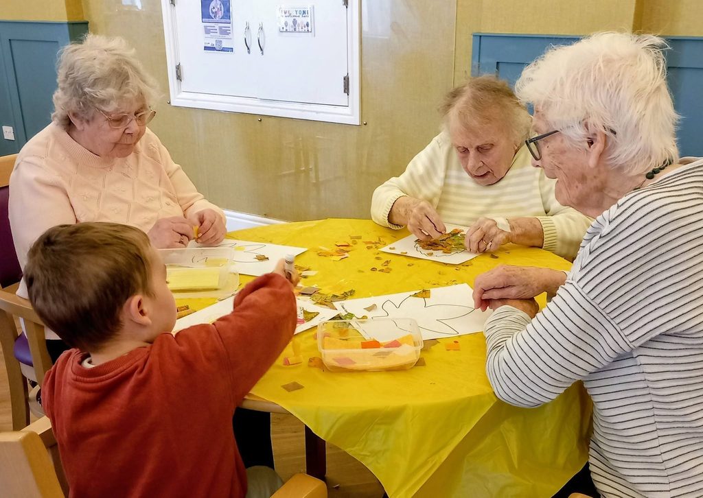 Three elderly women and a young boy sit around a round yellow table for morning activities, crafting with paper pieces on leaf-shaped outlines.
