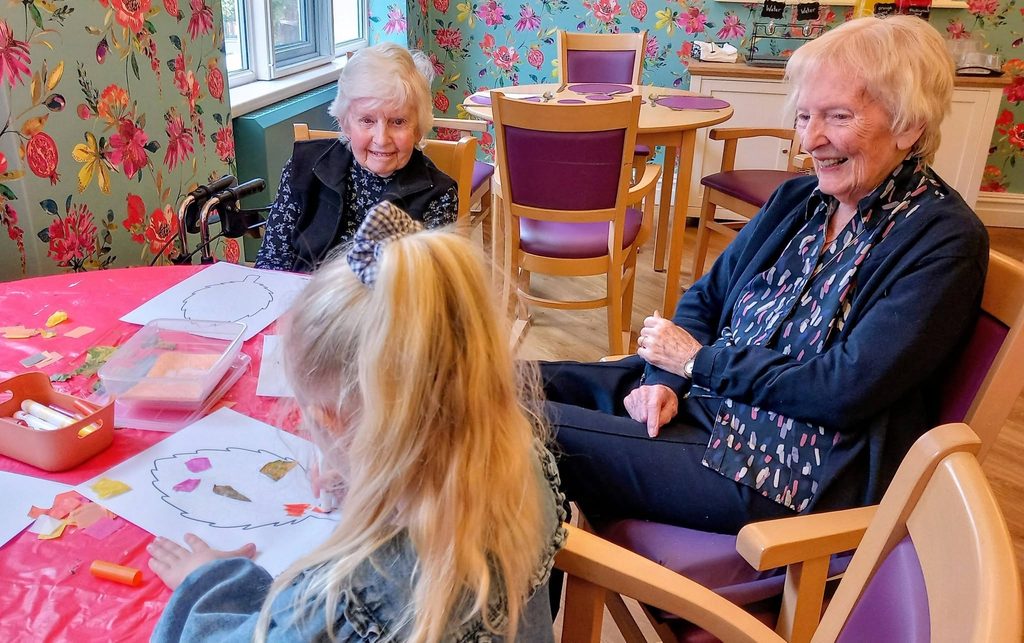 A young girl sits at a table enjoying morning activities as she does crafts with paper, watched by two elderly women.
