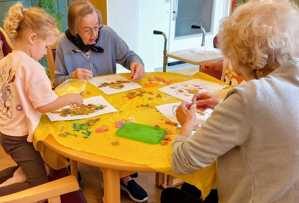 A young child and two elderly women sit around a round table covered in a yellow cloth, enjoying morning activities whilst sticking leaves onto paper crafts.