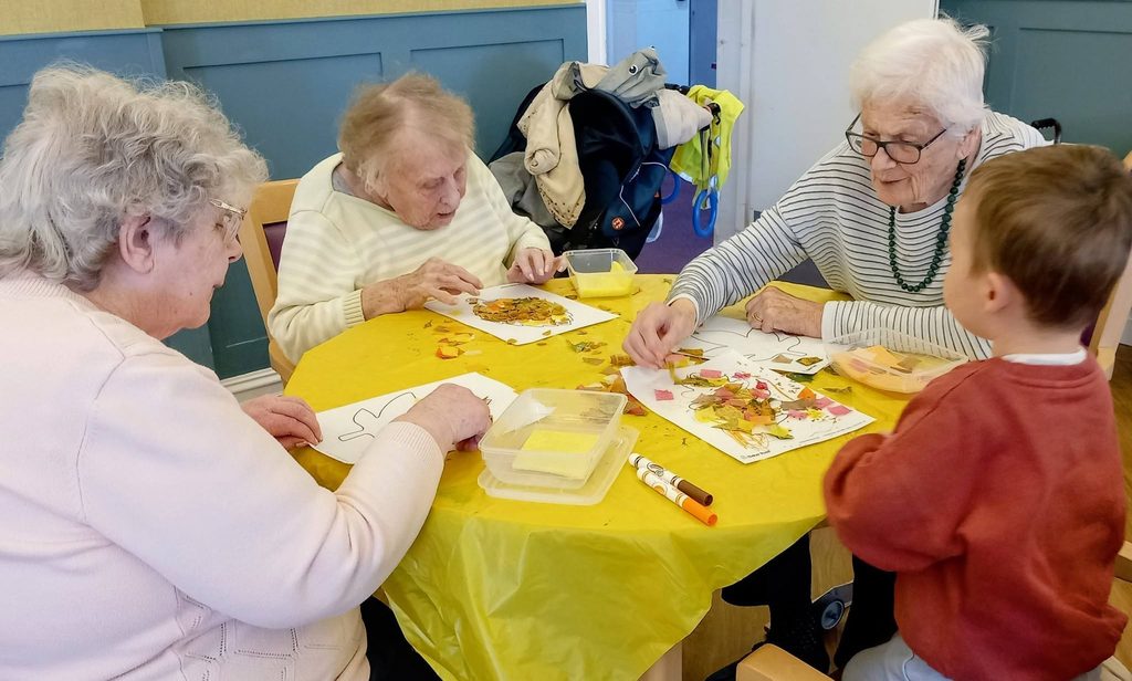 Three elderly women and a young child sit around a yellow-clothed table, engaged in morning activities as they glue leaves onto paper crafts.