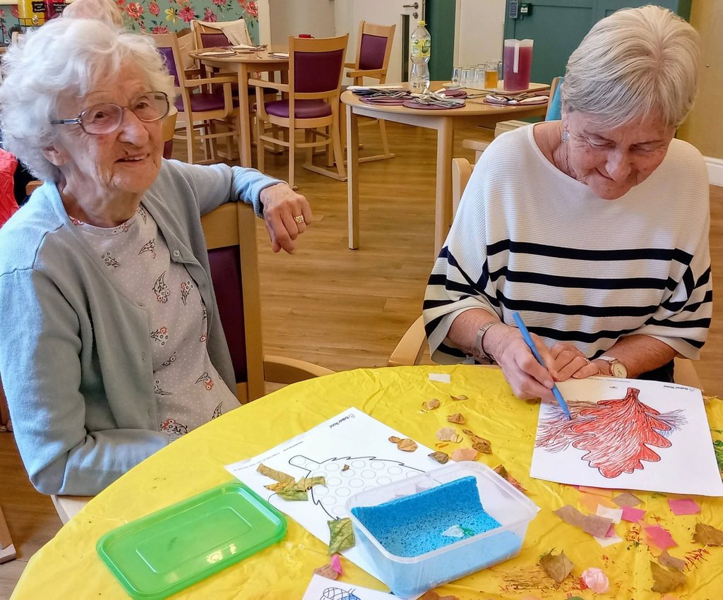 Two women sit at a yellow table, one smiling at the camera while the other enjoys morning activities by colouring in a picture of a tree.