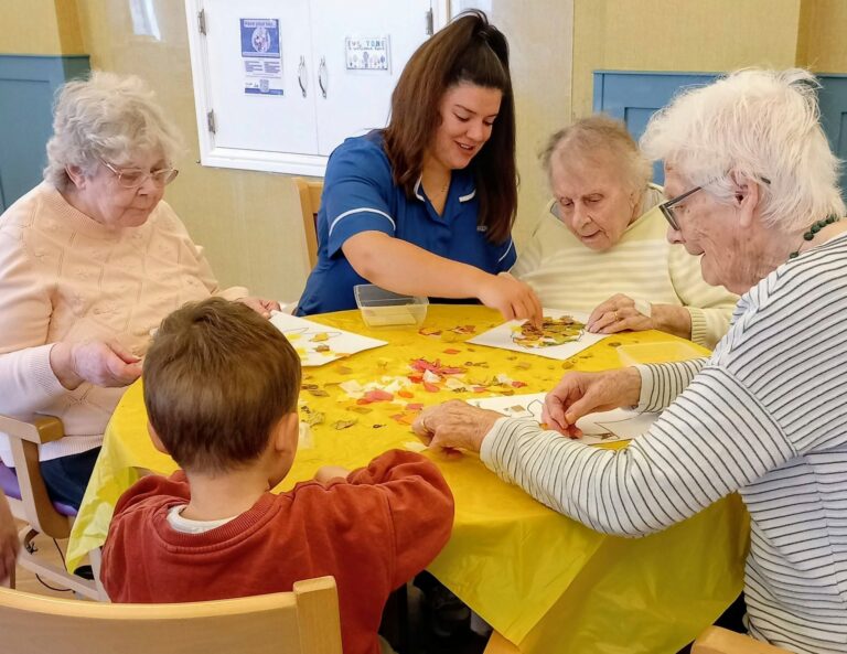 A care worker and a young boy join three elderly women for morning activities involving crafts at a yellow-covered table.