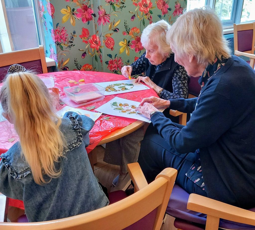 Two elderly women and a young girl sit at a table enjoying morning activities whilst creating leaf art collages.