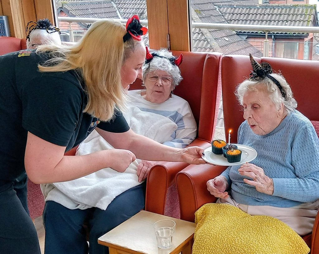 A woman offers a plate with two decorated fairy cakes and a lit candle to an elderly woman wearing a Hallowe’en headband, showcasing spooky fun and connected generations.
