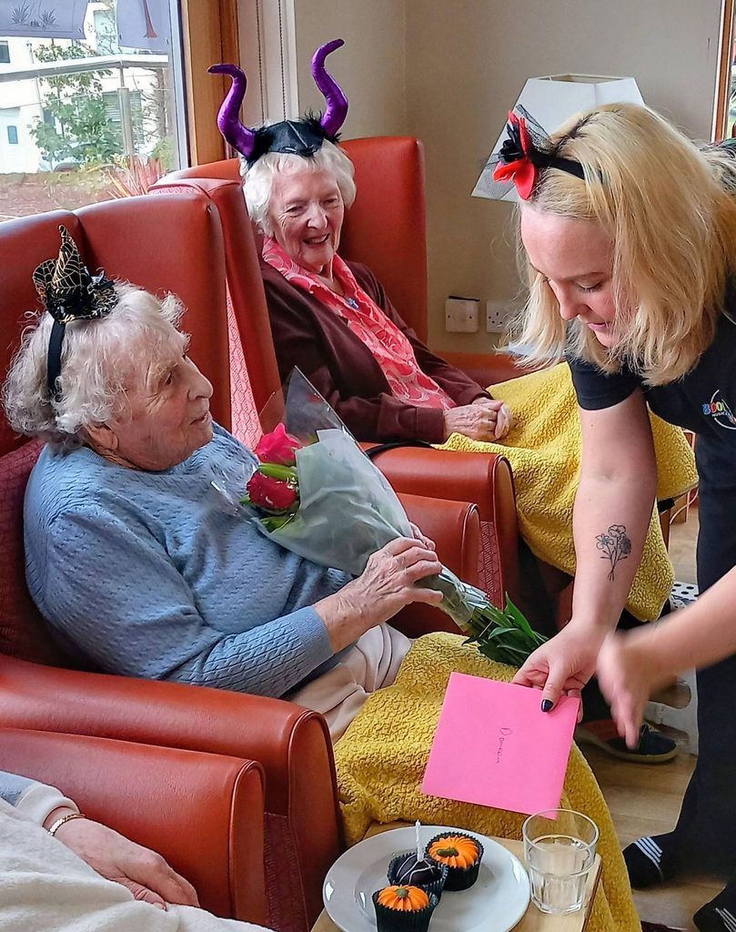 A woman in a black T-shirt hands a birthday smile on a pink card to an elderly woman in a blue jumper holding roses, whilst another woman wearing purple toy horns sits nearby in an armchair to share in the spooky fun.
