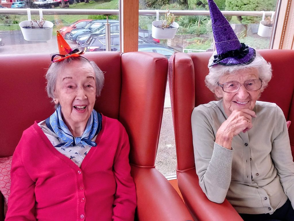 Two elderly women wearing small witch hats sit in red armchairs with birthday smiles.