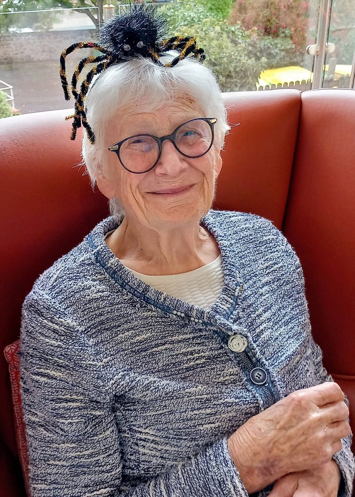 An elderly woman with white hair and spectacles wears a spooky spider hairband to celebrate her birthday.