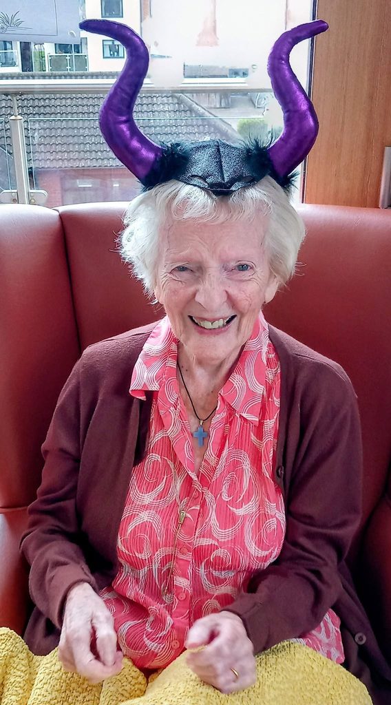 An elderly woman shares a birthday smile while wearing a headband with shiny purple devil horns.