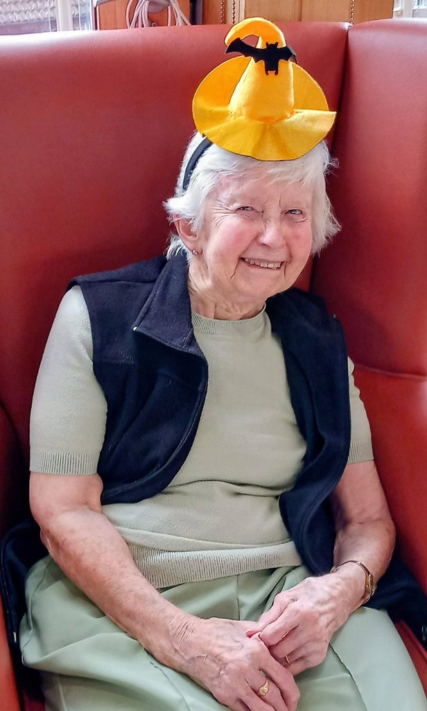 An elderly woman wearing a small orange Hallowe’en witch hat headband, smiling with spooky fun.