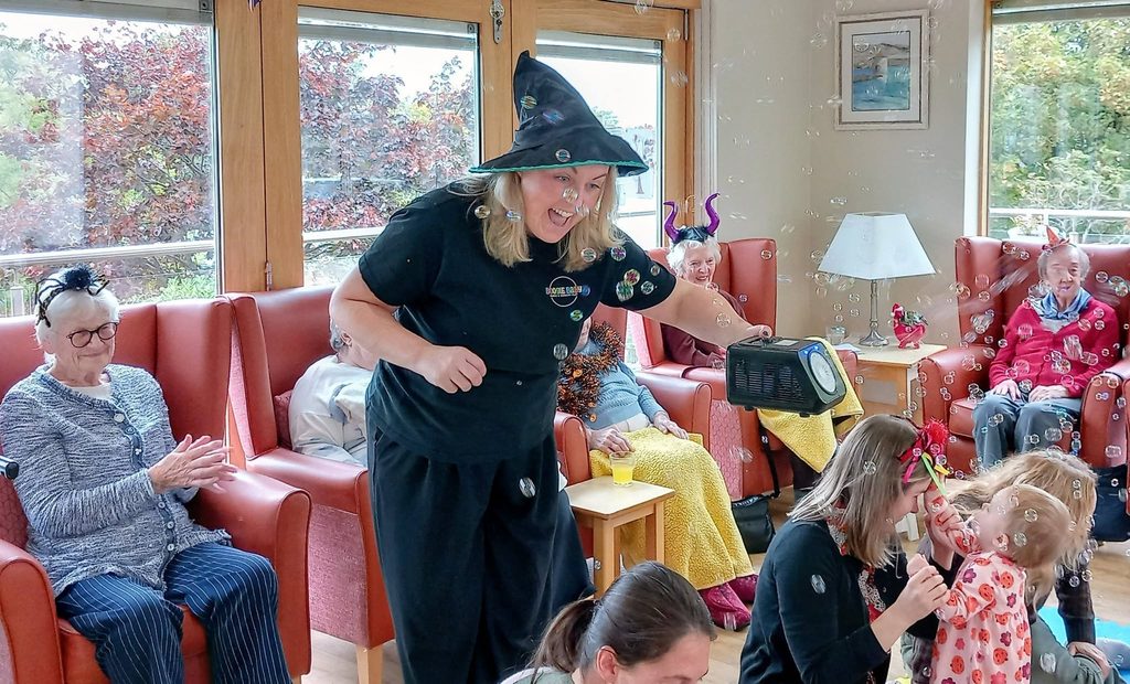 A woman wearing a witch’s hat blows bubbles with a machine for a group of elderly residents in a lounge, creating moments of connected generations and spooky fun.