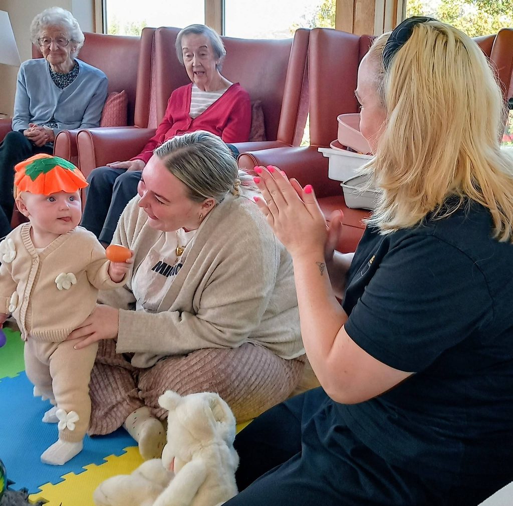 A woman helps a baby wearing a carrot hat stand on a play mat on an autumn morning while another woman applauds, watched by two older women sitting in armchairs as connected generations share the moment.