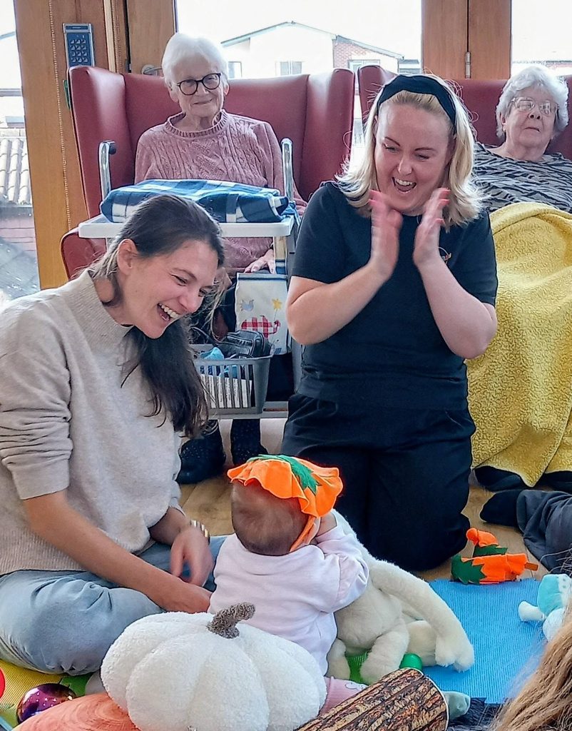 Two women sit on the floor playing with a baby wearing a pumpkin hat, while two older women look on from chairs in the background in this autumn photography family portrait.