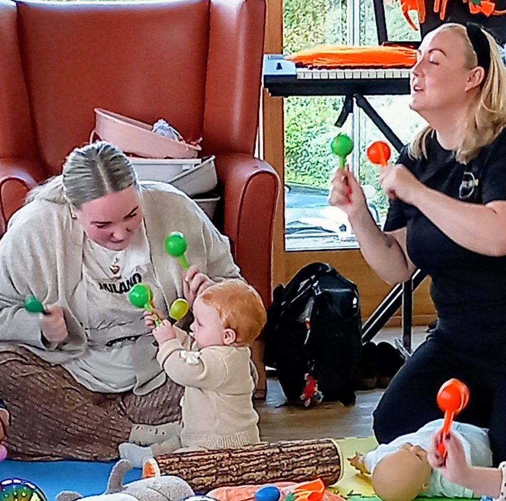 A woman and a young child sit on the floor playing with green maracas, while another woman sits nearby shaking an orange one during a connected generations autumn morning.