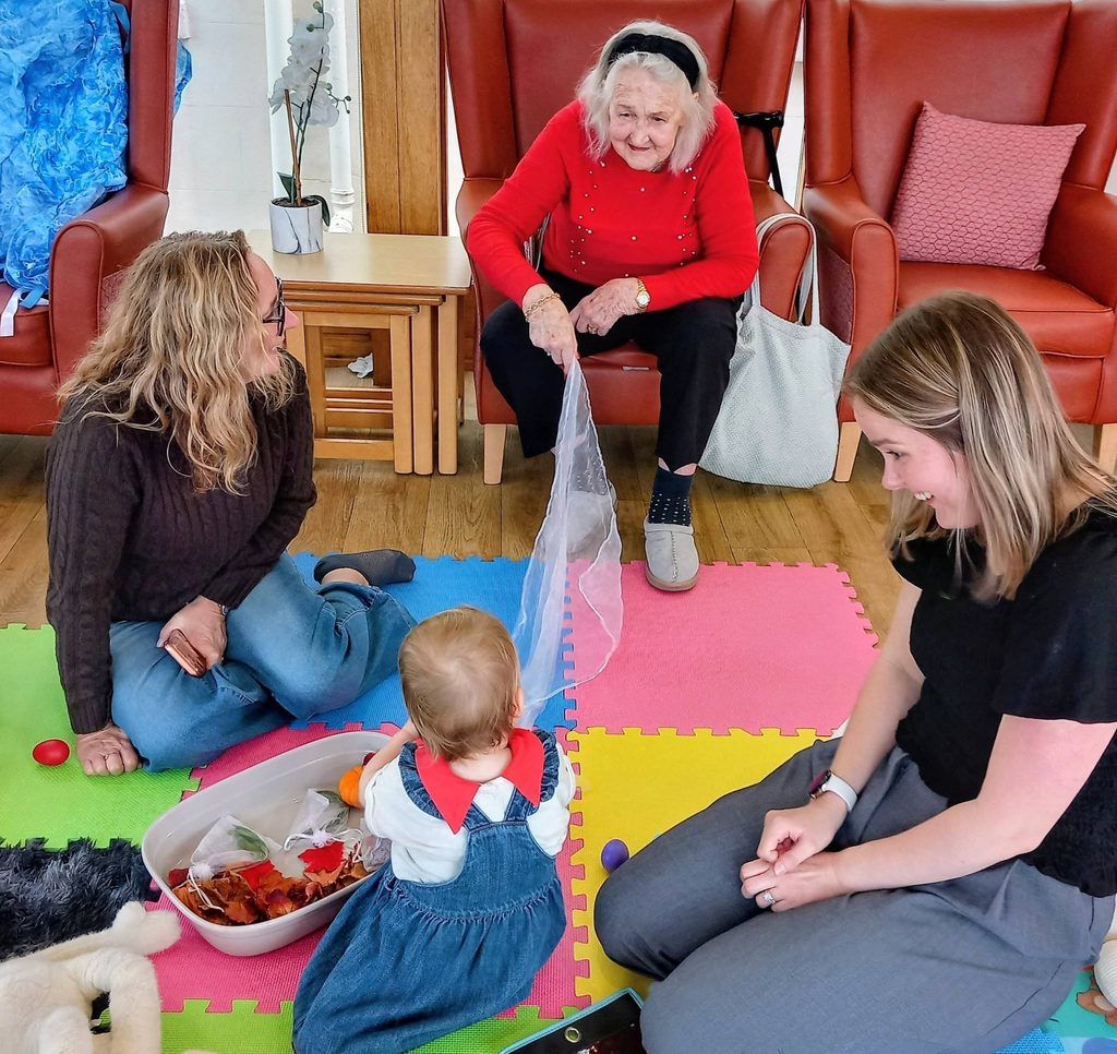 An elderly woman in a red jumper holds a sheer white fabric towards a toddler sitting on a colourful foam mat between two women in an autumn pumpkin patch.