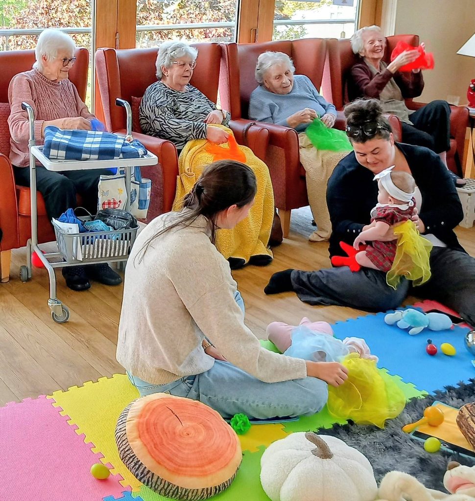 Four elderly women in armchairs and two women on a mat play with a baby using colourful fabric scraps in a cosy autumn setting.