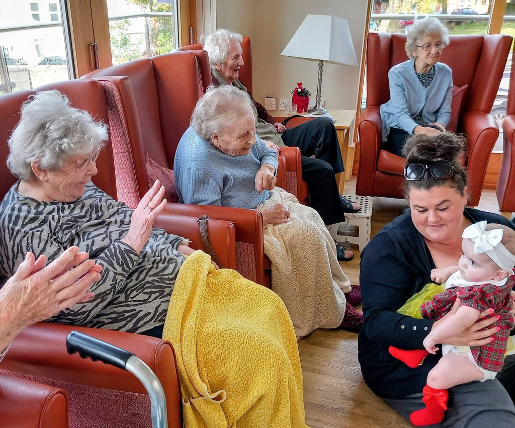 Four elderly women sit in armchairs alongside a woman holding a baby in a red tartan dress, capturing connected generations on an autumn morning.