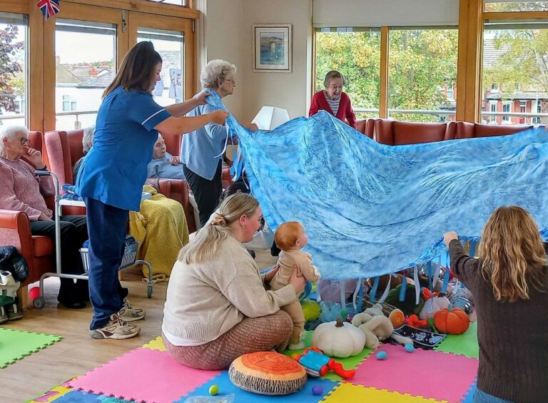 A carer and residents in a care home lift a large blue play parachute over a baby and autumn toys on a colourful floor mat.