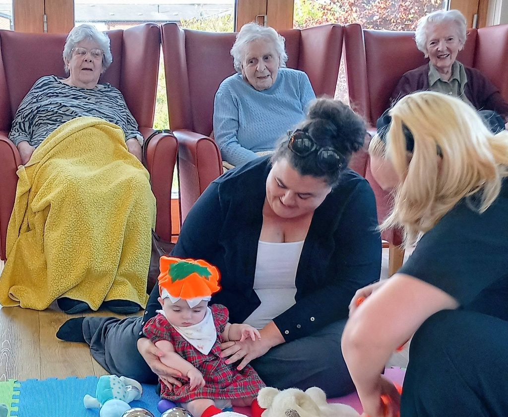 A baby in a red checked dress and pumpkin hat sits on the floor between two women during an autumn family gathering, while three elderly women watch from chairs in the background.