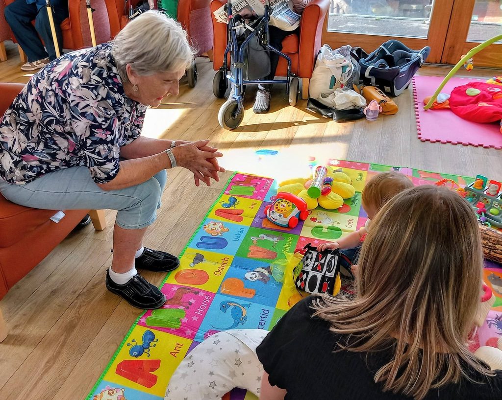 An elderly woman sits in a chair smiling at a toddler playing on an alphabet floor mat with an adult, capturing a moment of connected generations.